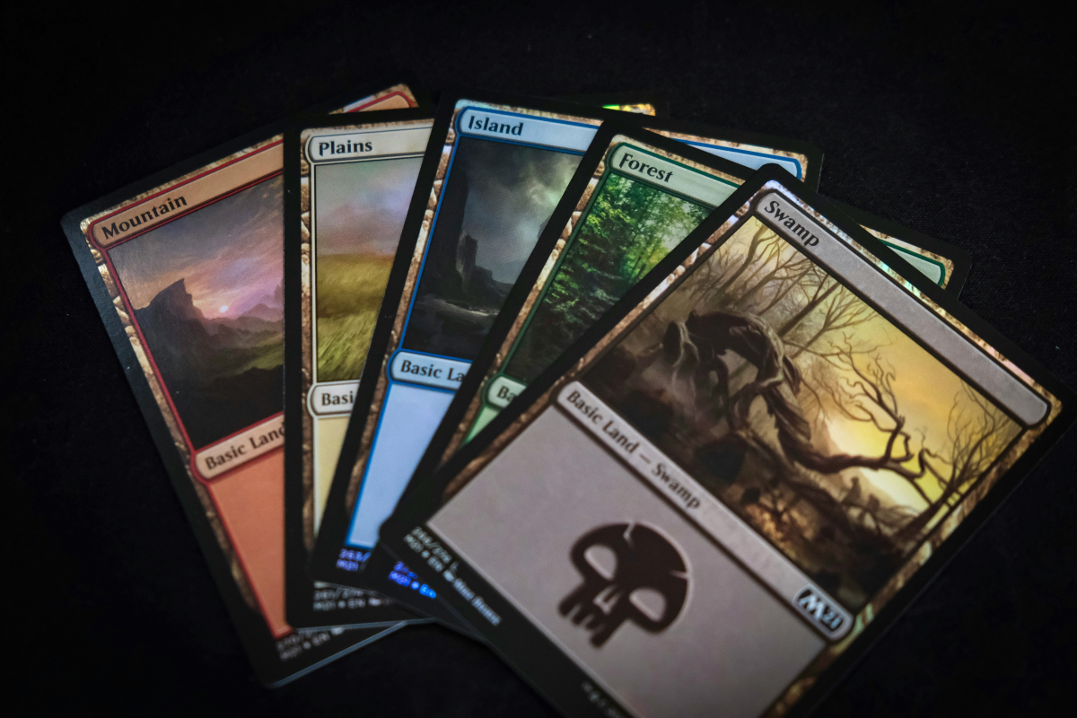 Shows an image of five different MTG basic lands spread out on a surface. This includes a Mountain, Plains, Island, Forest, and Swamp.